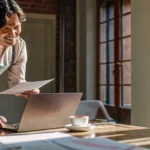 Small business owner reviewing financial documents on a laptop in a sunlit office